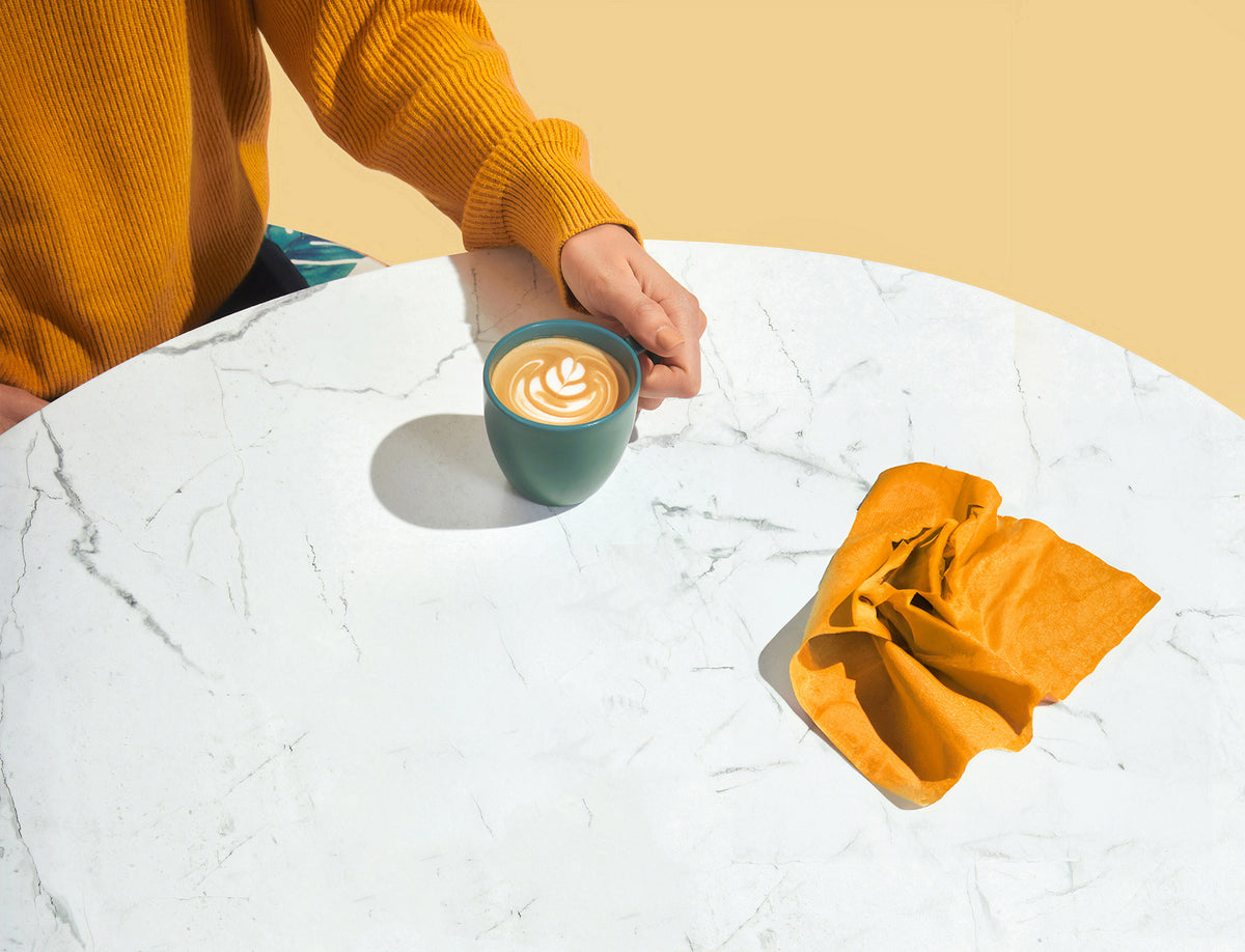 A person in a mustard yellow ribbed sweater holds a dark teal ceramic mug with latte art on a white marble table, next to a matching mustard yellow cloth against a solid yellow background.
